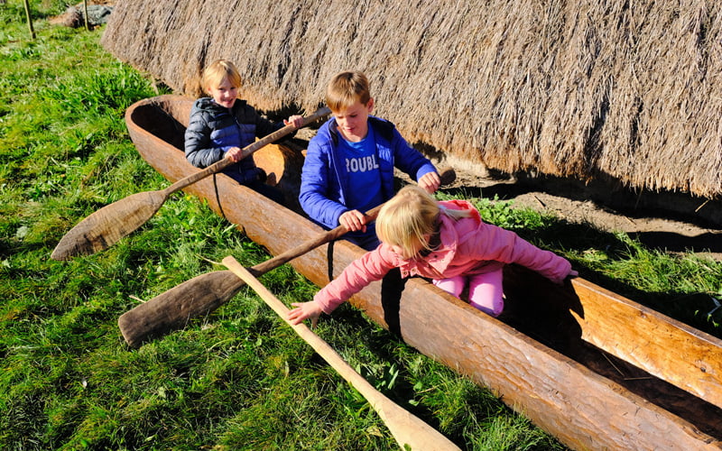 Drie kinderen met peddels in een boomstamkano op het gras naast een rieten dak